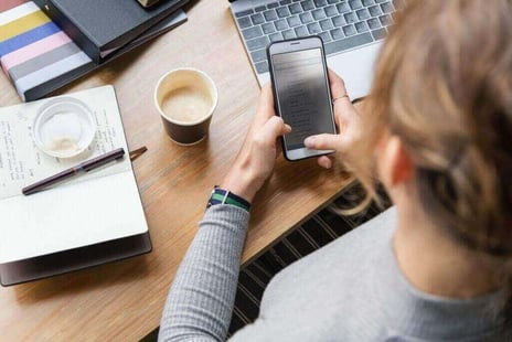 Woman sitting at a desk with a notepad, cup of coffee, and laptop. She is holding her cell phone in her hands.