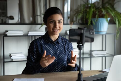 Woman sitting at a table, talking into a cell phone camera.